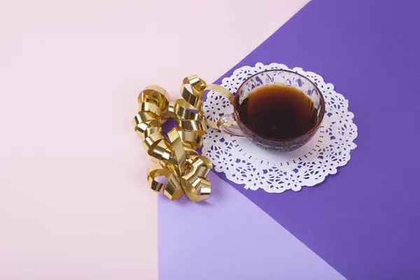 a violet cube made with colored paper to create a 3D optical illusion. On top is a gold Gift ribbon curled near a vintage pink coffee cup with a white lace doily. Vivid colors and minimal pop art photography.