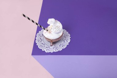 Üç boyutlu optik bir illüzyon yaratmak için renkli kağıttan yapılmış bir menekşe küpü. Üstte krem şanti ve beyaz dantelli altlığa serpiştirilmiş pembe bir fincan kahve var. Canlı renkler ve minimal pop sanat fotoğrafçılığı.