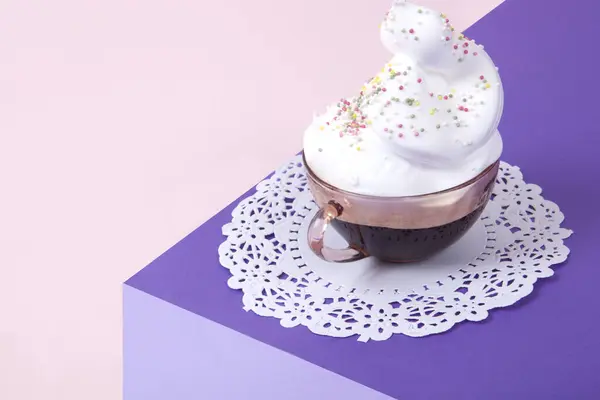 a  violet cube made with colored paper to create a 3D optical illusion. On top is a vintage pink cup of coffee with whipped cream and sprinkles on a white lace doily. Vivid colors and minimal pop art photography.