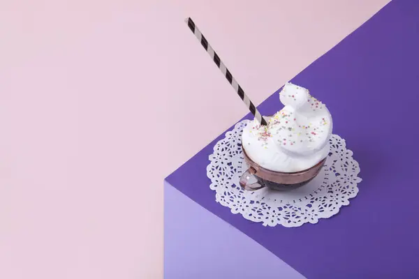 a  violet cube made with colored paper to create a 3D optical illusion. On top is a vintage pink cup of coffee with whipped cream and sprinkles on a white lace doily. Vivid colors and minimal pop art photography.