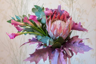 Renkli bir buket, ince zambaklar ve okaliptüs yapraklarıyla çevrili büyük bir proteyi sergiliyor. Yumuşak ışıklandırma düzenlemenin doğal güzelliğini arttırır.