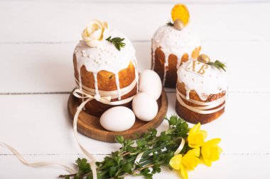 Hassas, hafif, modern tasarımı olan paskalya pastaları ve sarı nergisli beyaz ahşap arka planda yumurtalar. Paskalya konsepti. Geleneksel Paskalya yemekleri. Paskalya sembolü