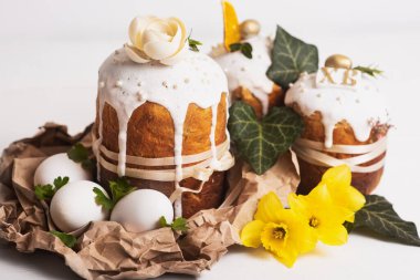 Hassas, hafif, modern tasarımı olan paskalya pastaları ve sarı nergisli beyaz ahşap arka planda yumurtalar. Paskalya konsepti. Geleneksel Paskalya yemekleri. Paskalya sembolü