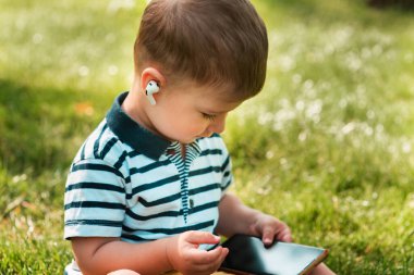 Yakışıklı Avrupalı bir çocuk yeşil çimlerin üzerinde kulaklığı kablosuz kulaklıkla akıllı telefon kullanıyor. Küçük bir çocuk alet kullanır. Bir çocuğun eğlence ve eğlence zamanı.