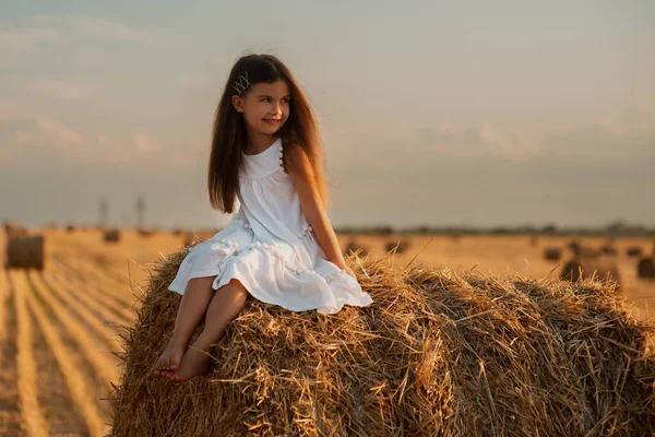 A cute little European-looking girl in a white summer dress sits on a bale of hay amidst a mowed field, on a farm. Summer time on a farm. Vacation on a ranch. Sunset in the village