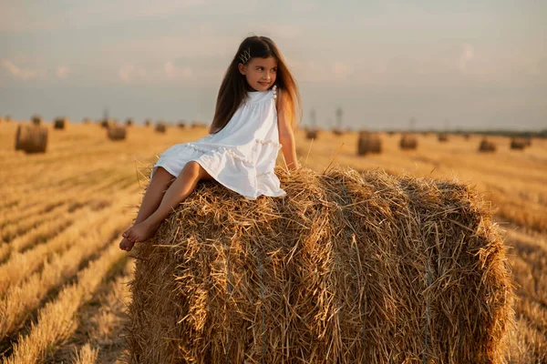 A cute little European-looking girl in a white summer dress sits on a bale of hay amidst a mowed field, on a farm. Summer time on a farm. Vacation on a ranch. Sunset in the village