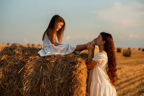 身穿白衣 长相可爱的欧洲小女孩坐在一捆干草上 与站在旁边的妈妈牵着手 在田野中央的一个农场里夏天在农场里母爱 — 图库照片