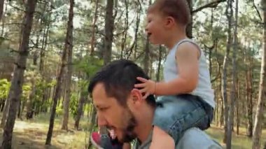 Baba, oğlu omuzlarındayken parkta yürüyor. Bir aile orman yolunda yürüyor. Mutlu bir çocukluk. Yüksek kaliteli FullHD görüntüler
