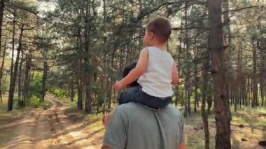 Baba, oğlu omuzlarındayken parkta yürüyor. Bir aile orman yolunda yürüyor. Mutlu bir çocukluk. Yüksek kaliteli FullHD görüntüler