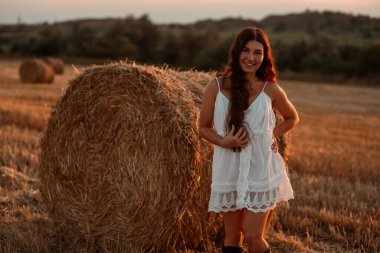 Beyaz saafanlı genç bir kadın gün batımında balyaların arasında buğday tarlasında vakit geçiriyor. Kırsal atmosfer, çiftlikte zaman. Yüksek kalite fotoğraf