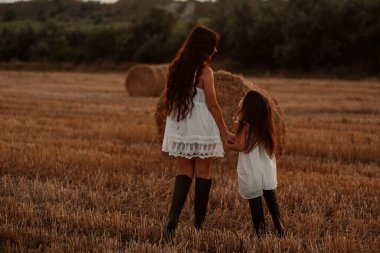 Beyaz saafanlarda bir anne ve küçük kızı gün batımında balyaların arasında buğday tarlasında vakit geçirirler. Kırsal atmosfer, çiftlikte zaman. Yüksek kalite fotoğraf