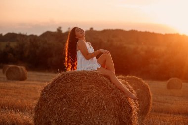 Beyaz saafanlı genç bir kadın gün batımında balyaların arasında buğday tarlasında vakit geçiriyor. Kırsal atmosfer, çiftlikte zaman. Yüksek kalite fotoğraf
