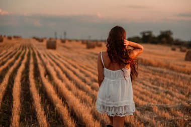 Beyaz saafanlı genç bir kadın gün batımında balyaların arasında buğday tarlasında vakit geçiriyor. Kırsal atmosfer, çiftlikte zaman. Yüksek kalite fotoğraf