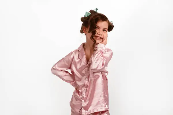 彼女の髪にカーラーと白い背景にピンクの絹のパジャマの緩いカールを持つかわいい少女は笑う 高品質の写真 ロイヤリティフリーのストック写真