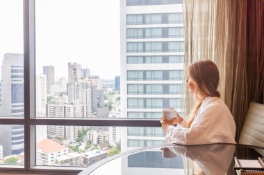 Modern bir otelde ya da gökdelen manzaralı bir dairede oturan bornozlu bir kadın. Lüks otellerde tatil. Otel odasında tatilde olan bir kadın.