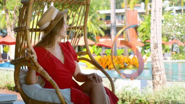 Yaz tatillerinde lüks bir otelde rakun koza salıncağında dinlenen güzel bir kadın turist. Hasır şapkalı ve kırmızı elbiseli romantik gezgin bayramlarda tropik tatil beldelerinde havuz başında dinleniyor..