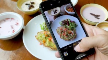 Erkek insan, modern akıllı telefonla sağlıklı kahvaltı yemeklerinin fotoğrafını çeker. Avokado tostu, yoğurt ve müsli. Yakın çekim..