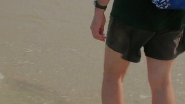 Gün batımında deniz kıyısında yalınayak yürüyen bir adam. Tayland, Phuket 'te yaz tatilindeki erkek turist. İnsan bacakları ve ayakları kumlu sahilde tek başına okyanusta su dalgalarında yürüyor..
