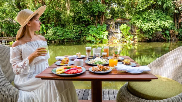 Girl relaxing and drinking healthy juice on breakfast or dinner on lake restaurant in luxury hotel. Woman in hat near green pond eating morning food and enjoy summer view. Concept of travel in Asia