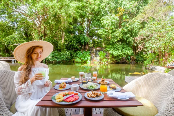 Girl relaxing and drinking healthy juice on breakfast or dinner on lake restaurant in luxury hotel. Woman in hat near green pond eating morning food and enjoy summer view. Concept of travel in Asia