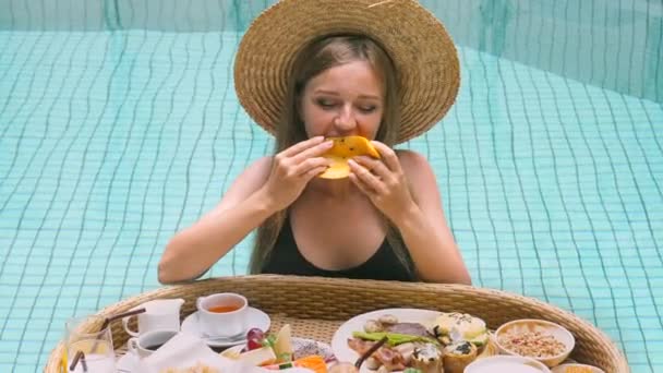 女人们戴着草帽在游泳池里 吃着漂浮的早餐盘里的美味芒果 享受热带水果 看着相机 豪华旅游 泰国度假 — 图库视频影像
