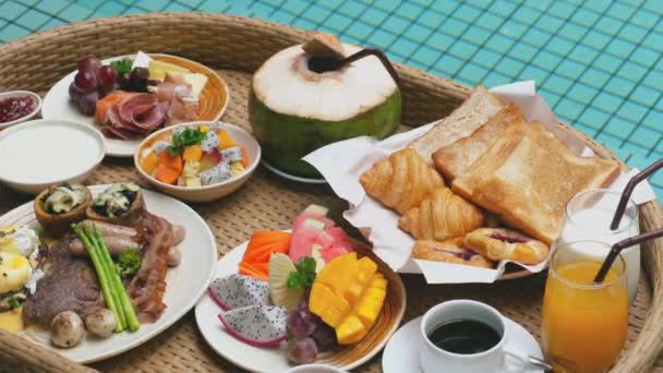 在带有游泳池的现代别墅的稻草托盘里享用美味的早餐 带着新鲜羊角面包 热带水果和果汁的漂浮早餐 在泰国的豪华旅行 — 图库视频影像
