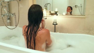 Beautiful young woman taking bubble bath at hotel or home, washing herself. Tub with foam bubbles. View from behind. Spa treatment, body care. Leisure time.