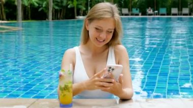 Happy smiling woman with mobile phone drinking tropical cocktail in swimming pool at luxury resort. Relaxed young traveler using smart phone surfing social media, checking news, playing mobile games.