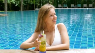 Beautiful woman in white swimsuit lounging in swimming pool, drink cocktail. Perfect for summer pool resort or spa hotel promotions. Portrait of smiling and relaxed female model enjoy tropical drink