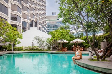 Woman enjoying luxurious tropical summer vacation. Female traveler sitting on edge of modern hotel swimming pool, surrounded by beautiful tropical landscape. Stylish architecture in background