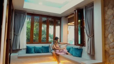 Couple in a beachfront resort, sitting together on couch, admiring sea view through window. Young family relaxed and comfortable, enjoying leisure time on vacation. Romantic getaways, beach vacations