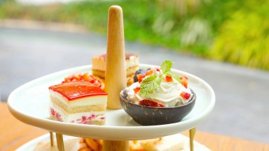 Elegance tea ceremony with shelf filled fresh pastries, cakes, and sandwiches. Afternoon tea, cafes, and food. The background with lush greenery. Sweet breakfast in cozy cafe