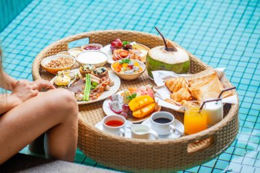 Floating breakfast tray in luxury poolside hotel. Woman on hotel breakfast with fresh croissants, tropical fruits and juices. Luxurious travel, resort vacation, summer holidays in Thailand