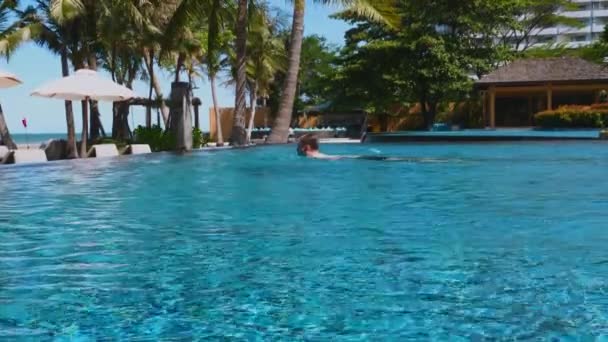 背景のヤシの木と海に囲まれた美しい青いスイミングプールでの男の水泳 リゾートでの熱帯の休暇や休日を促進するのに最適です — ストック動画