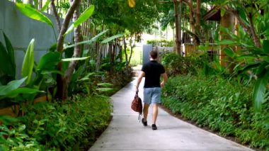 Male traveler with a backpack in his hands walking on villa in modern luxury tropical resort. Hotel in jungle. A man walks through the hotel among the greenery and trees. Back view