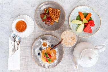 Outdoor served breakfast, includes glass of iced coffee, cup of tea, teapot, Belgian waffles, pancake with strawberry, plate of cut fruits. Perfect for hotel and sweet lunch concepts. Top view