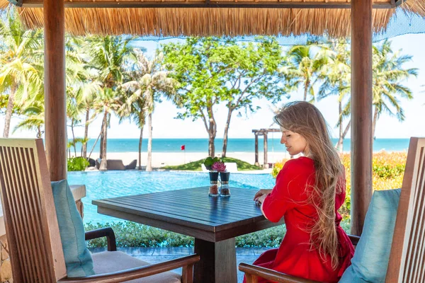 Woman on exotic summer vacation. Travel girl in red dress and hat sitting on breakfast at the table in hotel lounge near pool. Tropical beach restaurant with sea view