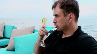 Angry unhappy alone man drinking alcohol cocktail on beachfront bar. Bad vacation. Sad man, displaying frustration, drinks an alcoholic cocktail at sea bar.