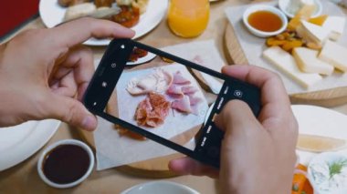 Male influencer and blogger captures breakfast with smartphone camera in restaurant. Close-up shots of a cold cuts for social media content. Brunch or lunch in hotel.