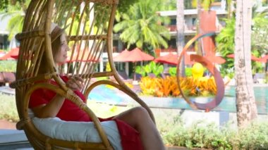 Young smiling woman lying in swing chair at poolside in tropical luxury hotel during her summer holidays. Romantic girl resting in hanging rattan cocoon in resort on vacations. Female tourist concept.