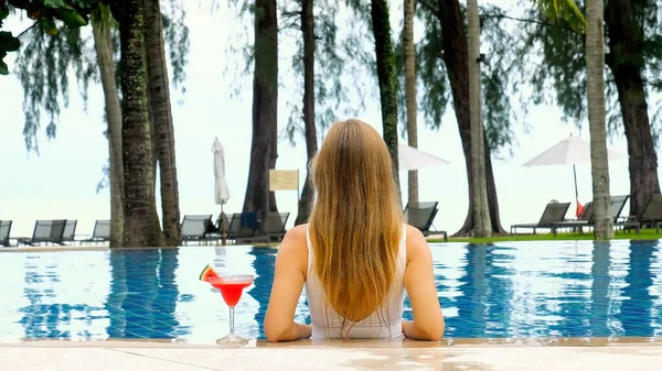 Back view of woman model, lounging in blue swimming pool in white bikini, tan during summer vacation. Relaxation at spa resort. Woman enjoying perfect beach holiday vacation at luxury beachfront hotel