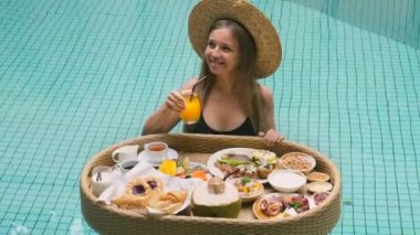 Smiling woman drinking fresh orange juice in swimming pool. Floating breakfast on tropical villa. Traveler tourist pool girl relaxing on vacation at luxury hotel, enjoy summer sunny day in Thailand