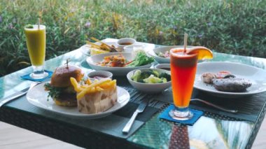 Table fool of various food - meat steak, beef burger, salad and vegetables, fresh fruit juices. Lunch or dinner on tropical vacation. Served table in luxury restaurant with greenery on background.