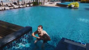 Male tourist plays in pool of luxury hotel. Man jumping in pool like sea star. Carefree vacation experience. Tropical setting and sea view in background. Travel and tourism, happy holidays.