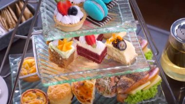 Close-up of delectable tier of macarons, scones, and petits cakes desserts, on a glass plate with a steaming teapot of green tea in background. Delicious sweet food.