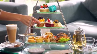 Woman hand delicately take a slice of cake from a towering display of sweet treats at a fancy restaurant afternoon tea, with cup of hot coffee and tea set nearby.