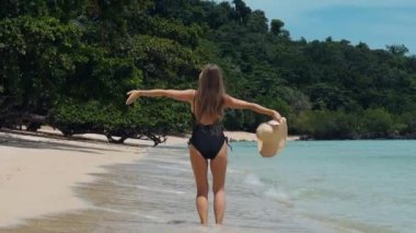 Happy young travel woman dancing on tropical sea beach, enjoy sunny day on travel holiday, fun and jumping. Back view of girl in swimwear celebrating summer vacation.