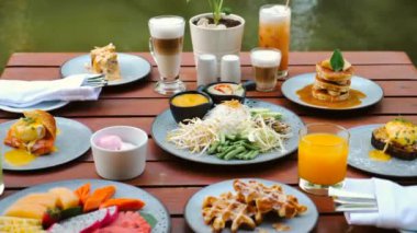 A luxury hotel breakfast with a table filled with fruits, pastries, eggs, juices, and coffee. The table is set near a green lake on a sunny day, highlighting the Asia travel concept.