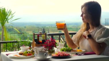 Woman on summer vacation enjoy fresh orange juice, sitting at table filled with breakfast food, surrounded by beautiful green valley view. Travel, resorts, and summer vacations