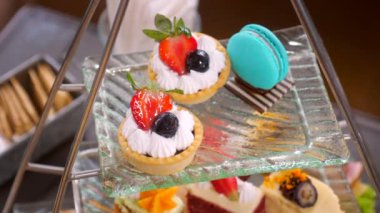 Woman chef hand put a bright blue macaron on afternoon tea stand with various colorful sweet desserts. Preparing delicious food for customers in restaurant or hotel. Close up.